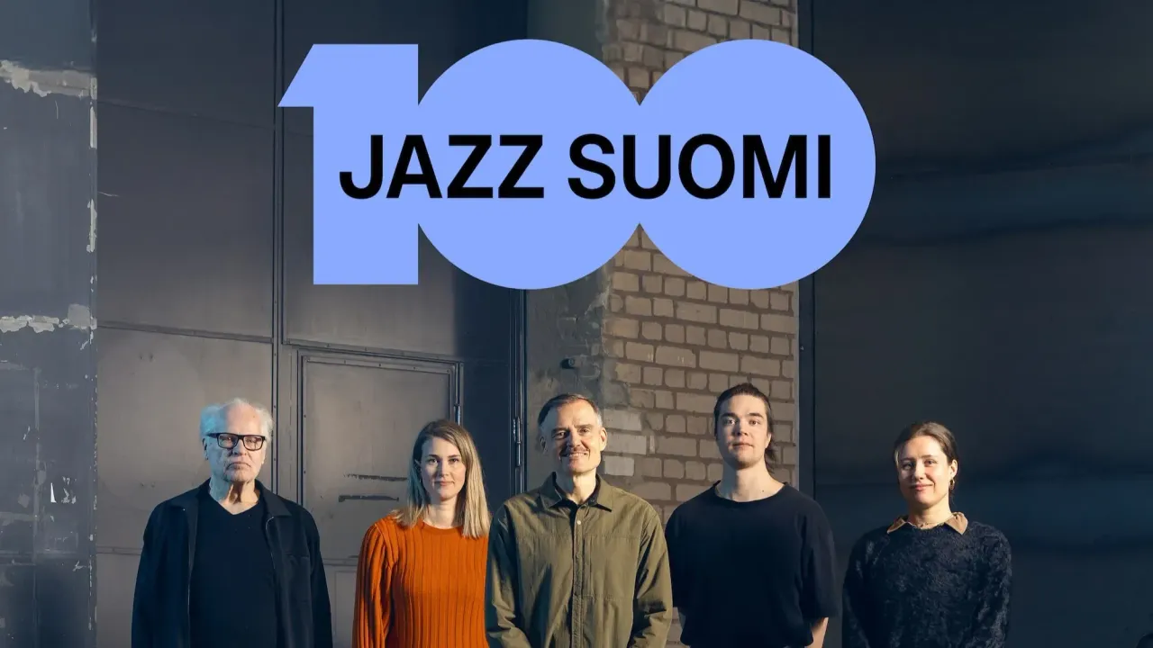 Viisi henkilöä seisoo rivissä teollisessa sisätilassa, ja yläpuolella on suuri vaaleansininen “100 JAZZ SUOMI” -teksti. Kuva näyttää juhlistavan Jazz Suomi -juhlavuotta.